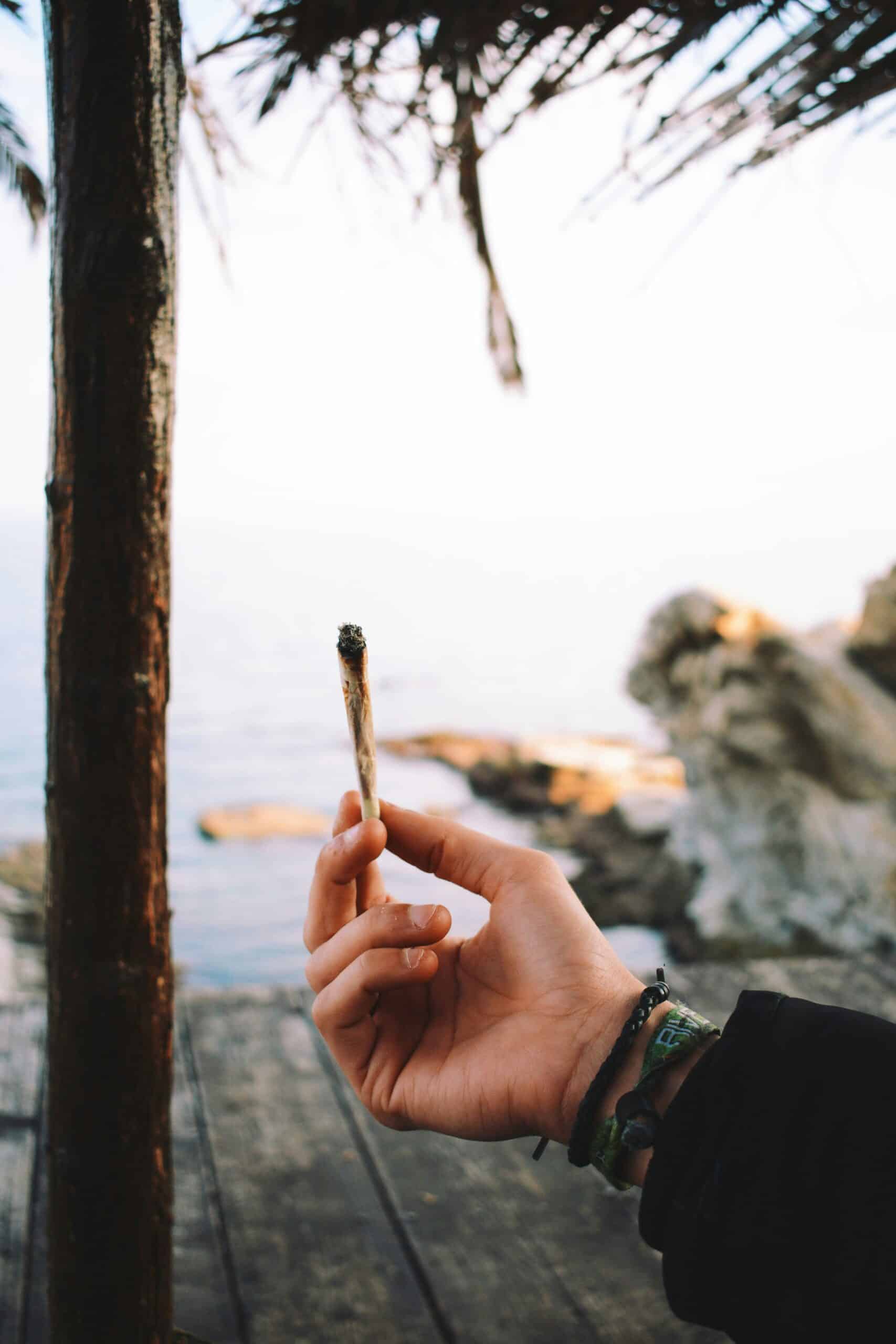 panos sakalakis dbdMjkC37yE unsplash A person holding a lit joint near a wooden deck overlooking the ocean, with a relaxed outdoor atmosphere. Image by Panos Sakalakis on Unsplash.com.