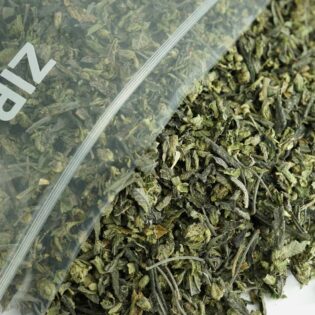 Bulk cannabis trim in a clear Zip-Zag bag with dried leaves, shake, and stems spilling onto a white surface.