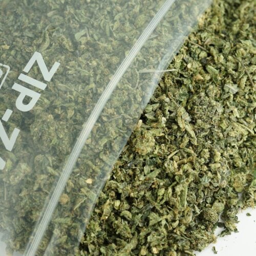 “Close-up of cannabis trim in a Zip-Zag bag with dried shake, leaves, and stems spilling onto a white surface.