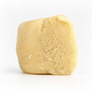 Close-up of a pale beige cube of cannabis extract on a crisp white background, showcasing a fine crumbly texture with subtle lighting and realistic surface detail.