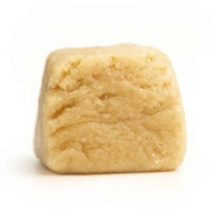 Close-up of a smooth, beige cube of cannabis concentrate with a slightly crumbly texture on a crisp white background, showcasing its soft, dense consistency and subtle sheen.