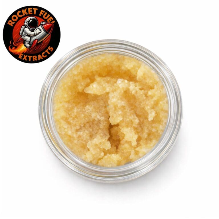 Top-down product image of cannabis concentrate in a clear glass jar with a golden, granular texture on a crisp white background, featuring the Rocket Fuel Extracts logo.