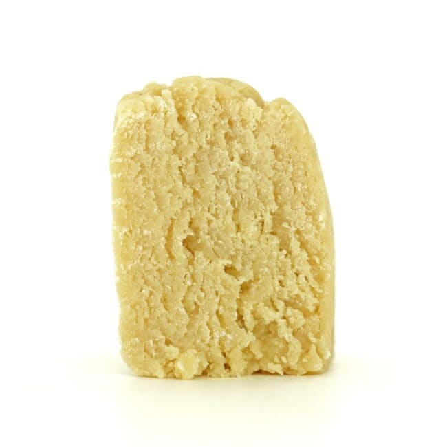Close-up of a light beige cannabis concentrate with a crumbly, textured surface on a white background.