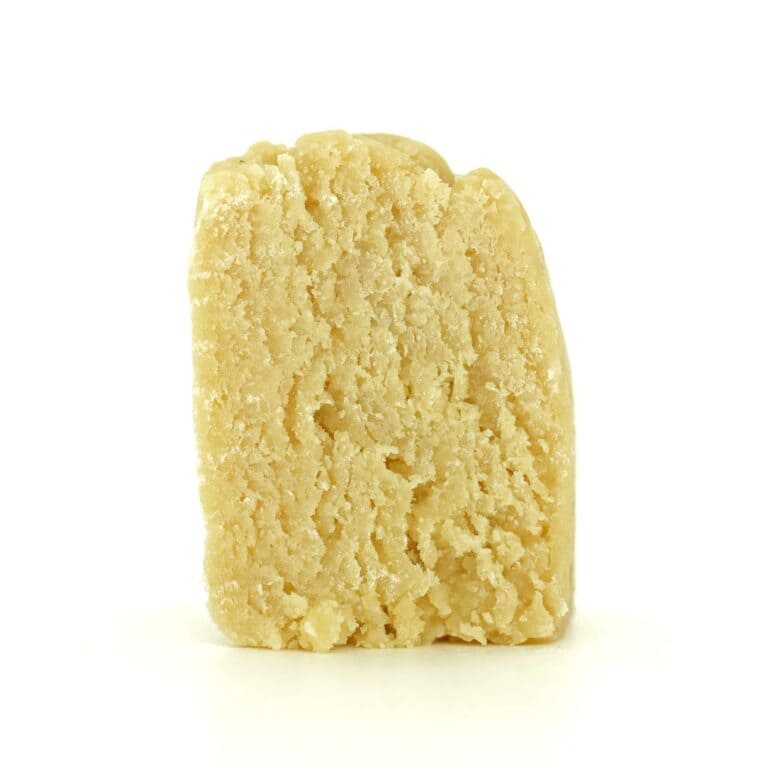 Close-up of a light beige cannabis concentrate with a crumbly, textured surface on a white background.