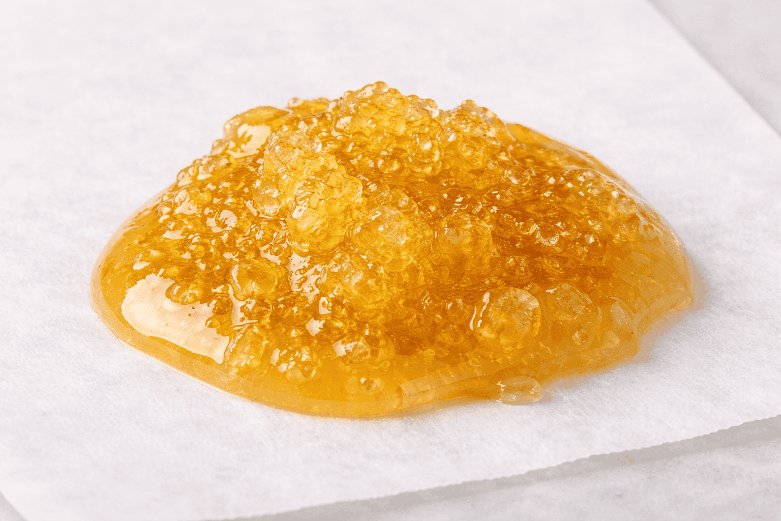 Close-up of glossy live resin with visible terpene-rich crystals and golden amber texture resting on white parchment against a clean light background