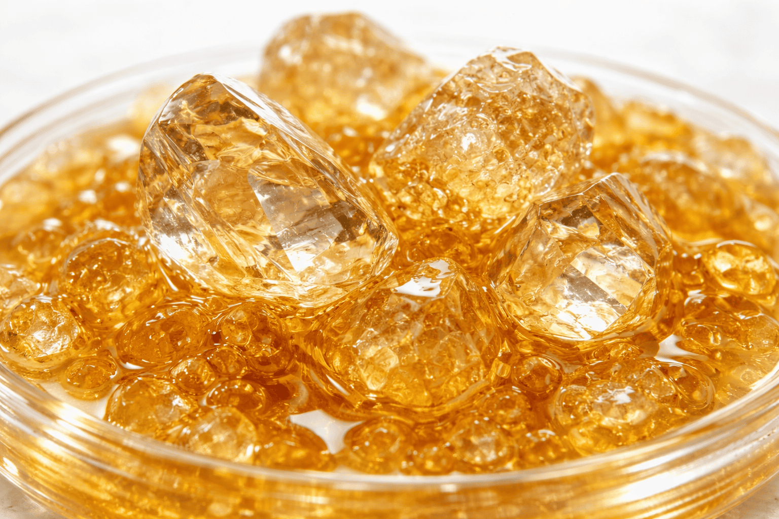 Clear THC diamonds with sharp crystalline facets resting in golden terpene sauce, displayed in a glass dish against a clean light background, highlighting clarity, texture, and glossy consistency.