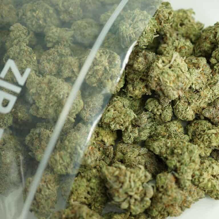 death star bulk close-up image of cannabis buds spilling out of a clear ZIP-ZAG plastic bag onto a white surface, showing dense green nugs with visible trichomes and orange hairs.