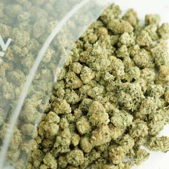 close-up image of bob cannabis buds spilling out of a clear ZIP-ZAG plastic bag onto a white surface, showing dense green nugs with visible trichomes and orange hairs.