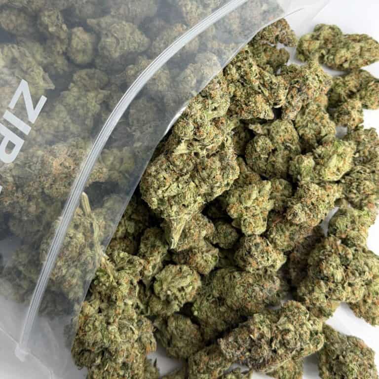 close-up image of cannabis buds spilling out of a clear ZIP-ZAG plastic bag onto a white surface, showing dense green nugs with visible trichomes and orange hairs.