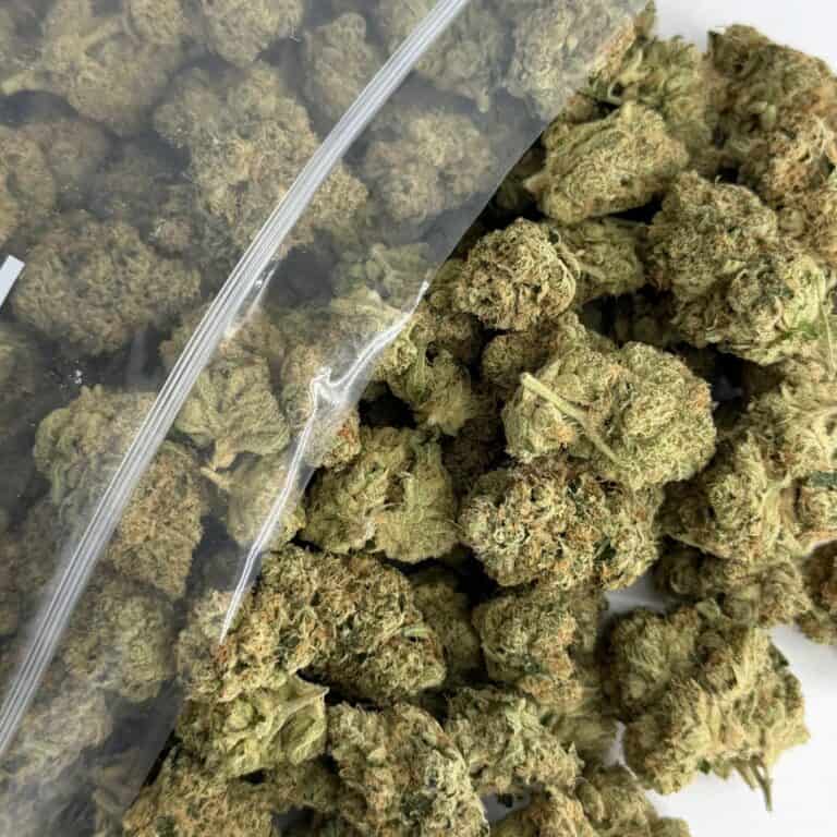 close-up image of cannabis buds spilling out of a clear ZIP-ZAG plastic bag onto a white surface, showing dense green nugs with visible trichomes and orange hairs.