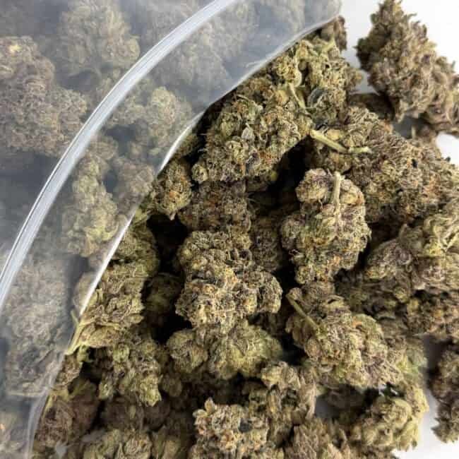 Gros plan sur des têtes de cannabis débordant d'un sachet en plastique transparent de marque ZIP-ZAG sur une surface blanche, montrant des têtes vertes denses avec des trichomes visibles et des poils orange.
