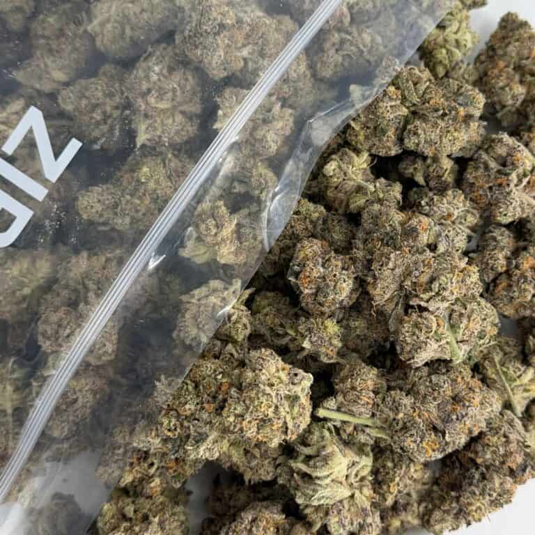 close-up image of cannabis buds spilling out of a clear ZIP-ZAG plastic bag onto a white surface, showing dense green nugs with visible trichomes and orange hairs.